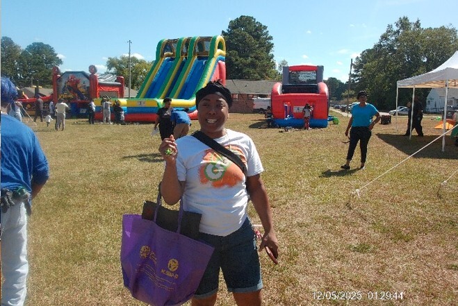 A woman holding a purple bag stands and smiles for a picture, with a inflatable slide in the background.